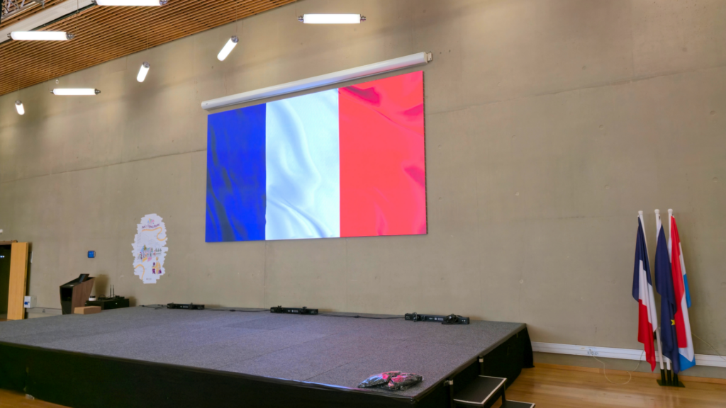 Grand écran LED de 5 mètres sur 2,75 mètres affichant le drapeau français, installé au lycée Vauban, au-dessus d'une scène. Plusieurs drapeaux sont placés sur la droite de la scène. écran QWARTZ 2.0 de 5m de longueur par 2m75 de hauteur au Vauban